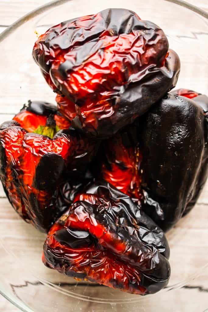 A close-up of several charred red bell peppers with blistered, blackened skins, ideal for making Roasted Red Pepper Soup, piled in a glass bowl on a light wooden surface.