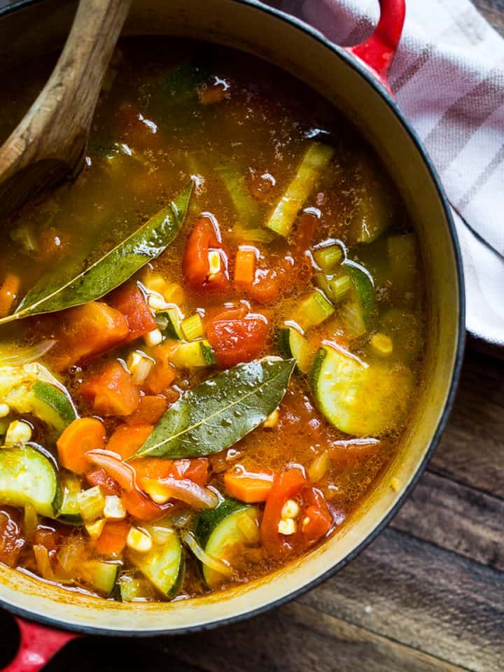A red pot filled with Late Summer Vegetable Soup (with Basil Pistou), brimming with zucchini, tomatoes, carrots, corn, celery, and bay leaves. A wooden spoon rests inside and a white towel lies nearby on a wooden surface.