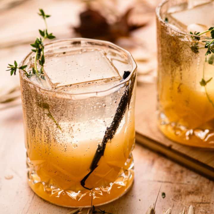 Two lowball glasses with vanilla pear mocktail and a large ice cube, vanilla bean pod. The glass is garnished with fresh thyme.