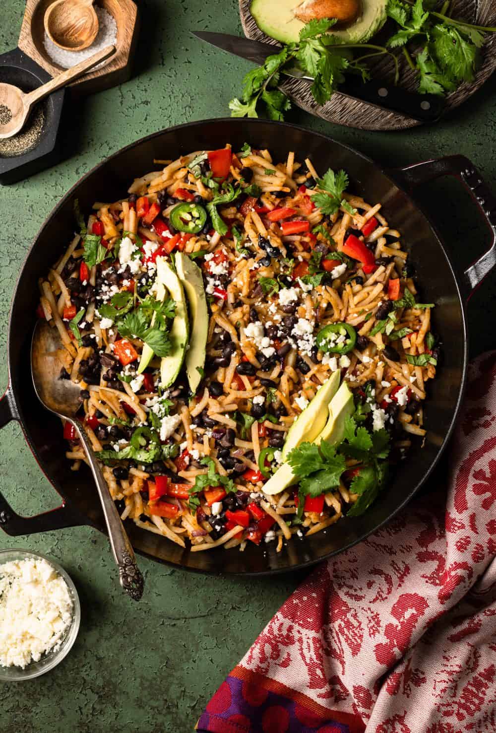 Cooked potato hash in a pan topped with avocado, the addition of cheese, herbs and a colorful napkin, small bowl of cheese, herbs and salt and pepper on the side.