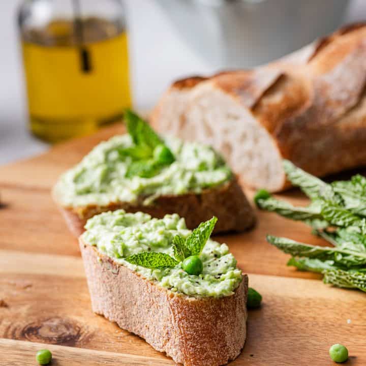 Slices of crusty bread topped with a creamy green spread, garnished with fresh mint leaves and peas, are arranged on a wooden cutting board. A loaf of bread and a bowl of pea spread and olive oil are visible in the background.