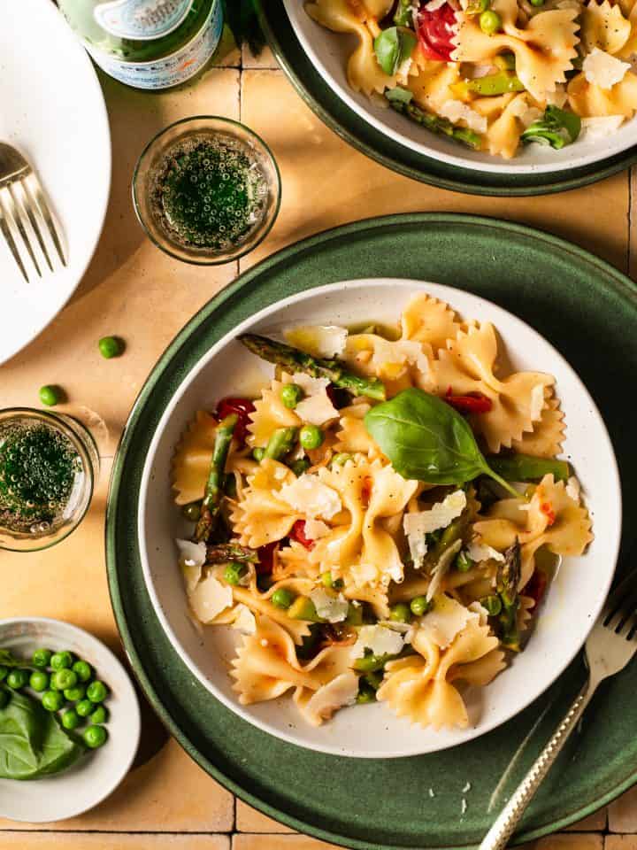 A bowl of farfalle pasta with vegetables and herbs on a green plate. In the background, theres a bottle of sparkling water, a glass, and more plates of pasta on a wooden table. The setting is warm and inviting.