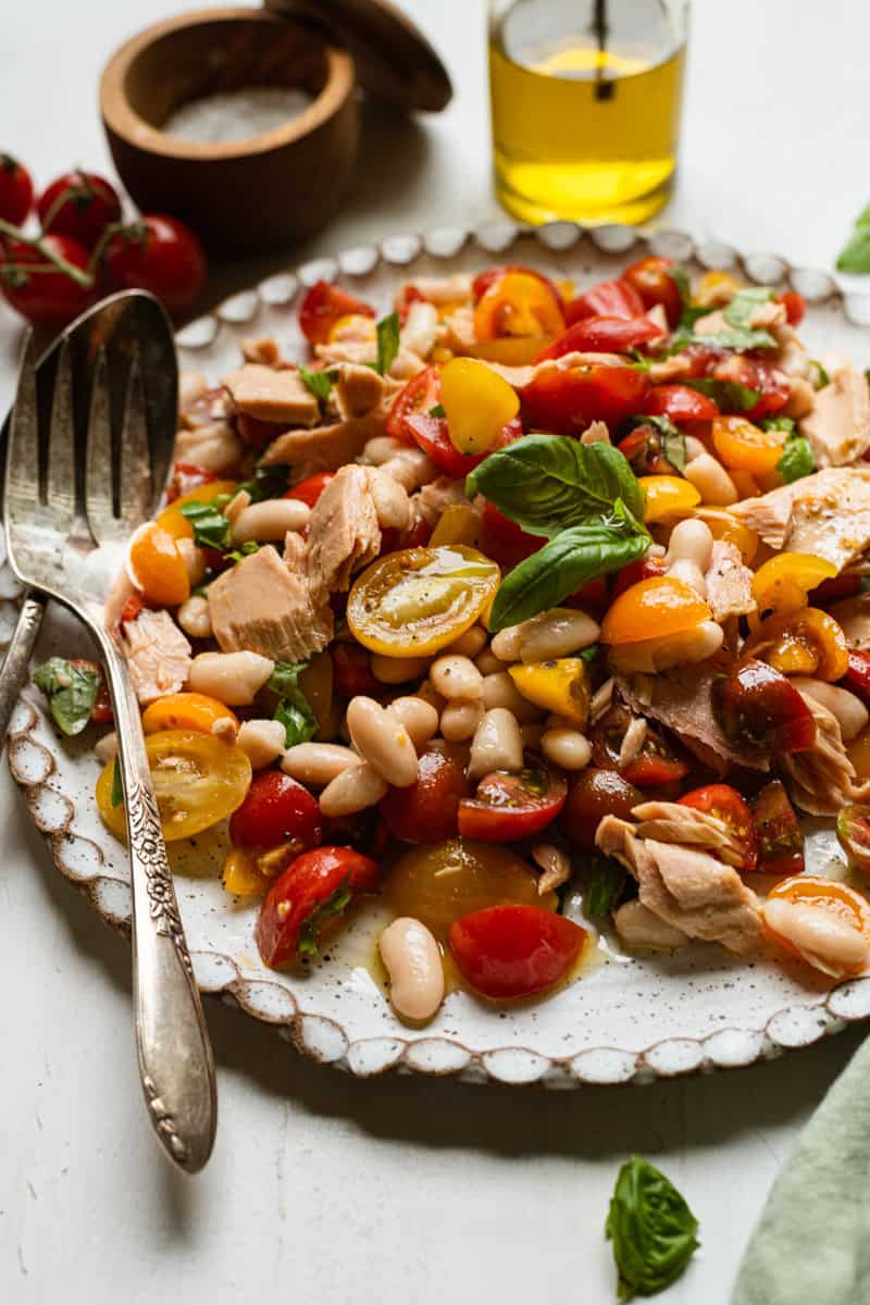 A plate of salad with colorful cherry tomatoes, white beans, chunks of tuna, and fresh basil leaves, served with a metal fork and spoon. A bottle of olive oil and a wooden bowl are in the background.